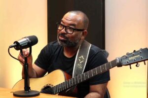 Pastor Luiz Arcanjo cantando e falando ao microfone enquanto toca violão durante gravação em estúdio.