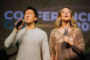 Pastora Jackie Hayashi e Lucas Hayashi cantando em momento de louvor durante conferência cristã, com microfones nas mãos e expressão de adoração.