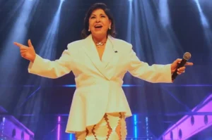 Roberta Miranda se apresenta no palco com traje branco, cantando e segurando o microfone sob iluminação cênica.