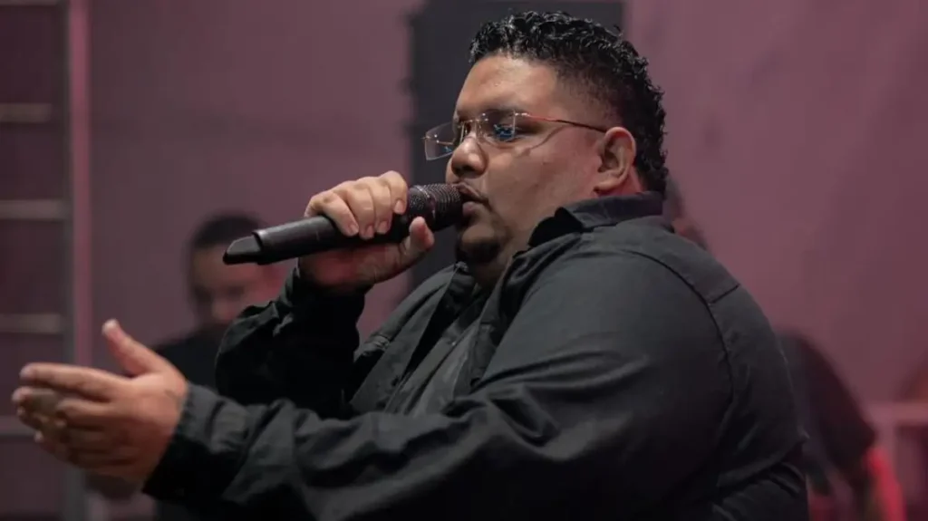 Israel Santos canta ao microfone durante apresentação em palco com iluminação rosada ao fundo