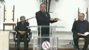 pastor Edson Cursino prega em púlpito da Assembleia de Deus Ministério de Taubaté durante culto com líderes sentados ao fundo