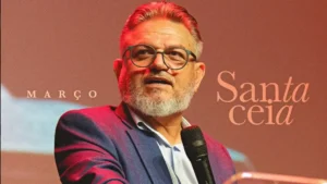 Pastor Francisco Júnior Romão fala ao microfone em evento religioso com telão “Santa Ceia” ao fundo