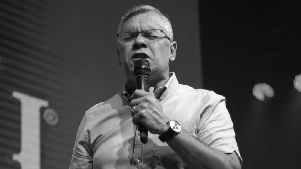 Pastor Jairo Fernandes, da Manah Church, fala ao microfone durante culto em palco, em foto em preto e branco