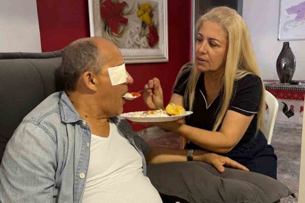Cantora Andrea Fontes alimenta o esposo sentado em poltrona, com curativo nos olhos