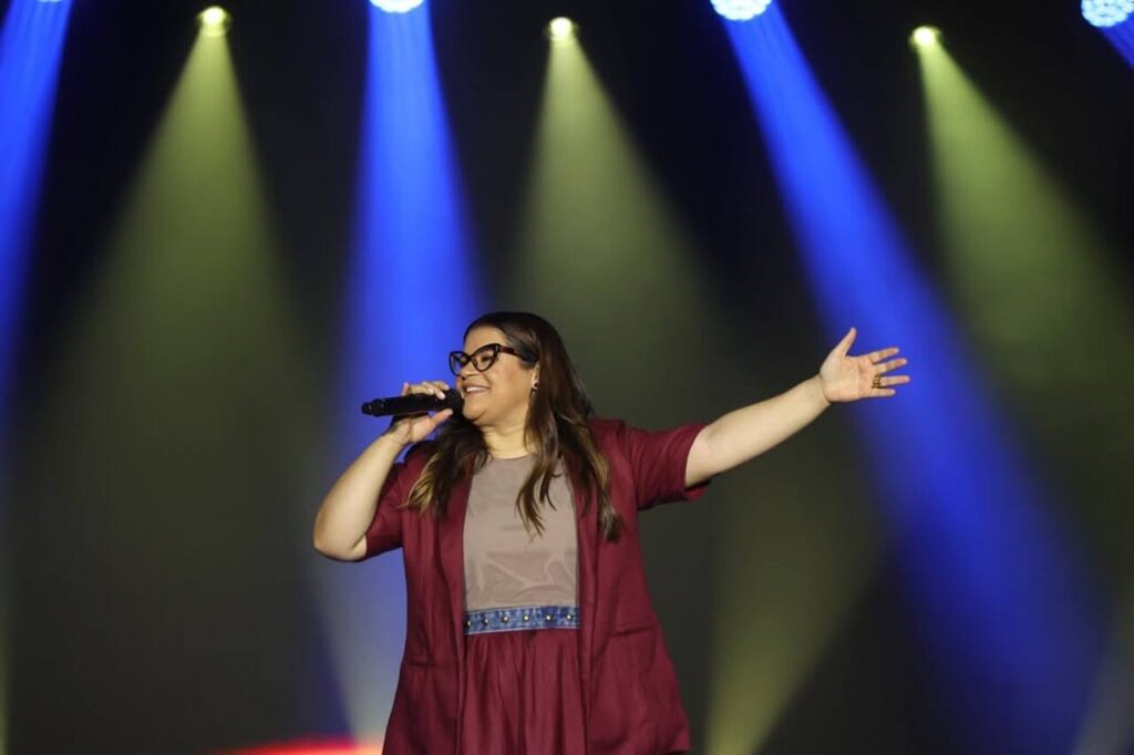 Cantora Midian Lima ministra louvor em palco, segurando microfone e com o braço estendido, sob iluminação colorida durante apresentação gospel.