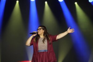Cantora Midian Lima ministra louvor em palco, segurando microfone e com o braço estendido, sob iluminação colorida durante apresentação gospel.