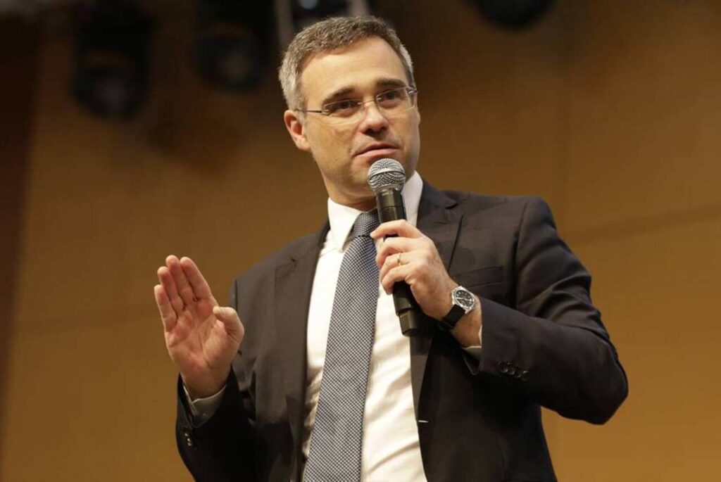 Ministro André Mendonça discursa segurando microfone, usando terno escuro e gravata, durante evento em ambiente fechado.