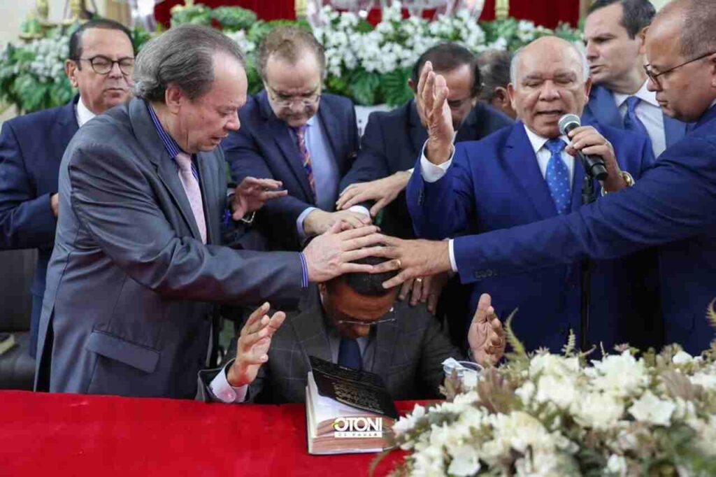 Otoni de Paula recebe oração com imposição de mãos durante culto evangélico, enquanto líderes religiosos oram ao seu redor.