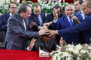 Otoni de Paula recebe oração com imposição de mãos durante culto evangélico, enquanto líderes religiosos oram ao seu redor.