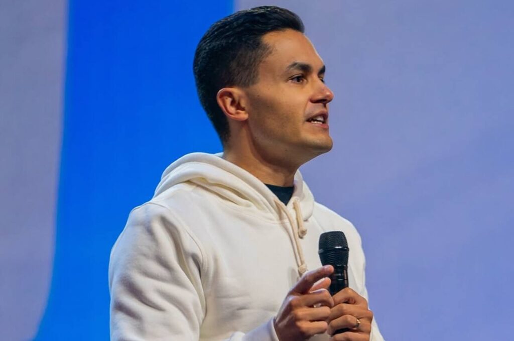 Davi Miranda Neto segurando microfone durante ministração, vestindo moletom claro, em ambiente de igreja ou conferência cristã.