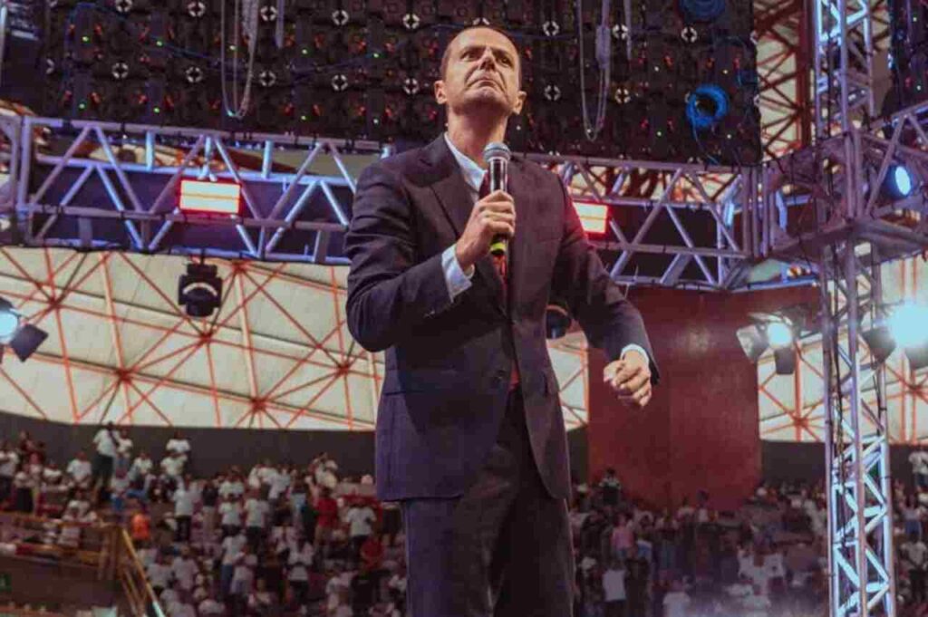 Pastor Juliano Fraga em palco de grande evento, segurando microfone, com público ao fundo e estrutura de iluminação profissional.