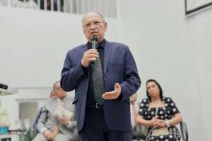 Pastor Napoleão Falcão fala ao microfone durante culto em igreja, usando terno escuro, com fiéis sentados ao fundo.