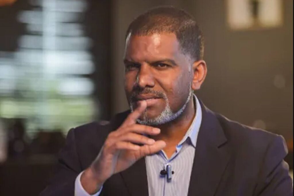 Pastor Oliver Costa em ambiente interno, usando blazer escuro, com a mão próxima ao rosto, em expressão reflexiva durante entrevista ou gravação.