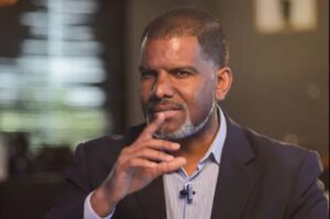 Pastor Oliver Costa em ambiente interno, usando blazer escuro, com a mão próxima ao rosto, em expressão reflexiva durante entrevista ou gravação.