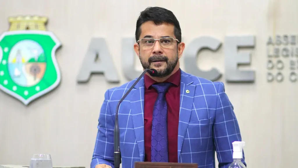 apostolo luiz henrique fala ao microfone em plenaria com a logo da alece ao fundo