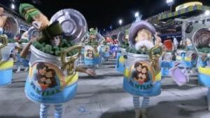 integrantes de escola de samba desfilam fantasiados de lata de conserva com caricaturas e plateia ao fundo no sambodromo