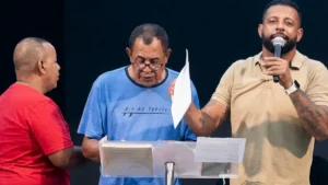 pastor fala ao microfone no altar enquanto homem lê documentos em púlpito durante culto na iptm