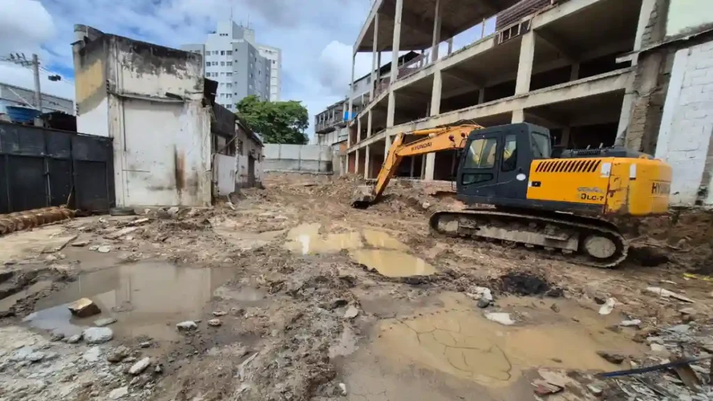 Escavadeira em terreno lamacento com poças de água e estrutura de prédio em construção ao fundo, em obra de casa de apoio para idosos