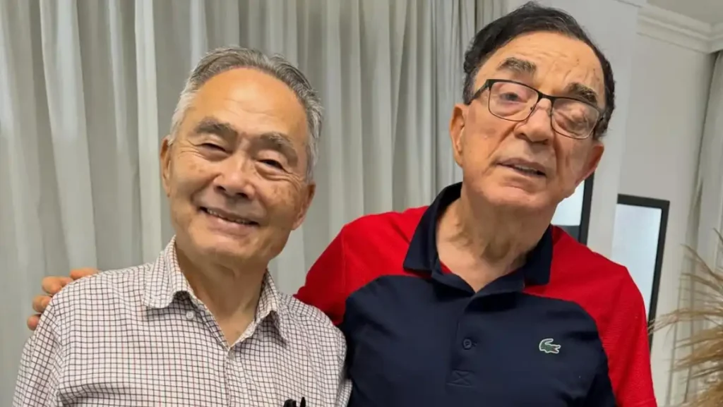 Ouriel de Jesus posa ao lado de Hidekazu Takayama dentro de uma residência, com cortinas claras ao fundo; os dois aparecem sorrindo para a câmera.