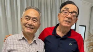 Ouriel de Jesus posa ao lado de Hidekazu Takayama dentro de uma residência, com cortinas claras ao fundo; os dois aparecem sorrindo para a câmera.