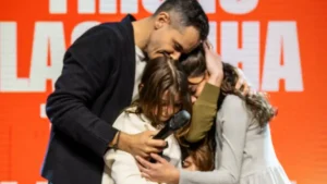 Pastor Paulo Oliveira abraça Juju Murrieta Oliveira e uma jovem no palco enquanto segura um microfone durante anúncio de renúncia na Igreja Batista da Lagoinha em Miami