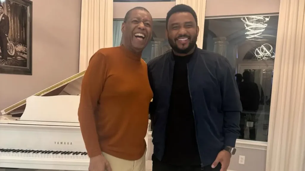 Ron Kenoly posa ao lado do cantor Eli Soares em sala com piano branco e cortinas claras