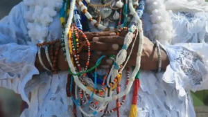 Pessoa com mãos cruzadas usa traje branco rendado e colares de contas coloridas em contexto de ritual religioso