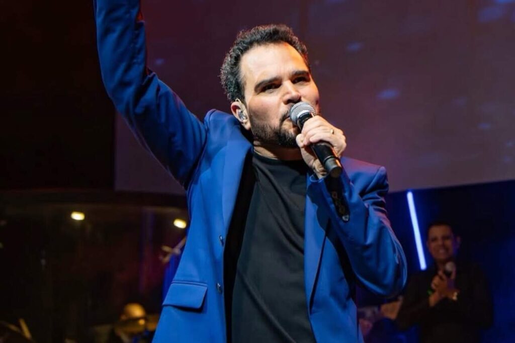 Cantor Luciano Camargo cantando ao microfone com a mão levantada durante apresentação em evento gospel