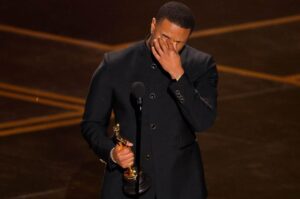 Ator emocionado segurando estatueta do Oscar durante discurso no palco da premiação.