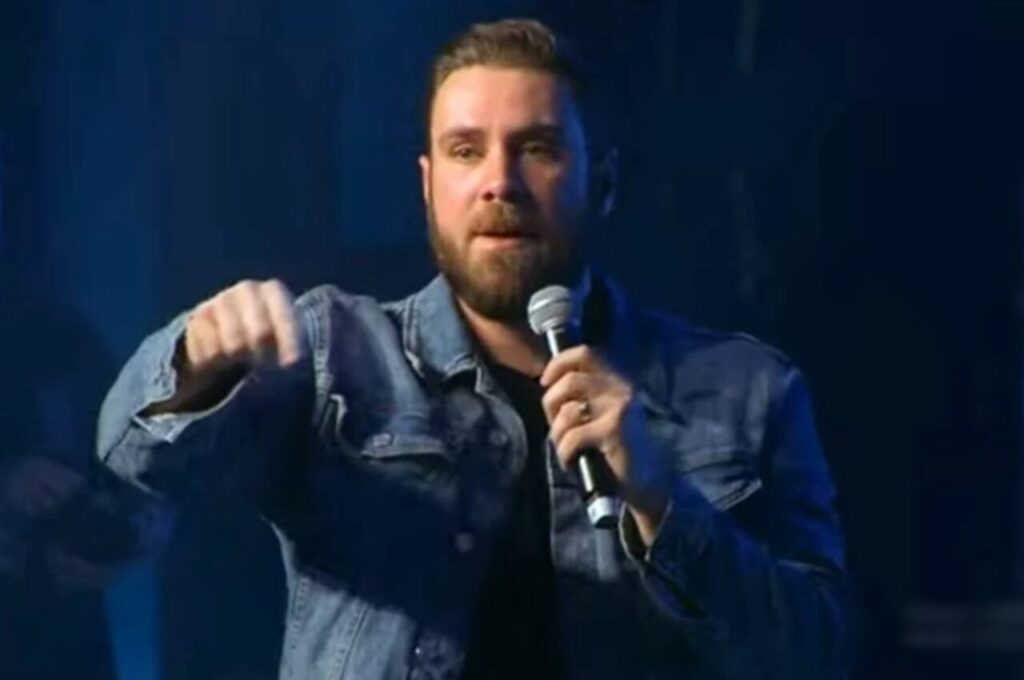 Pastor André Fernandes pregando com microfone em mãos durante culto, fazendo gesto de ênfase.