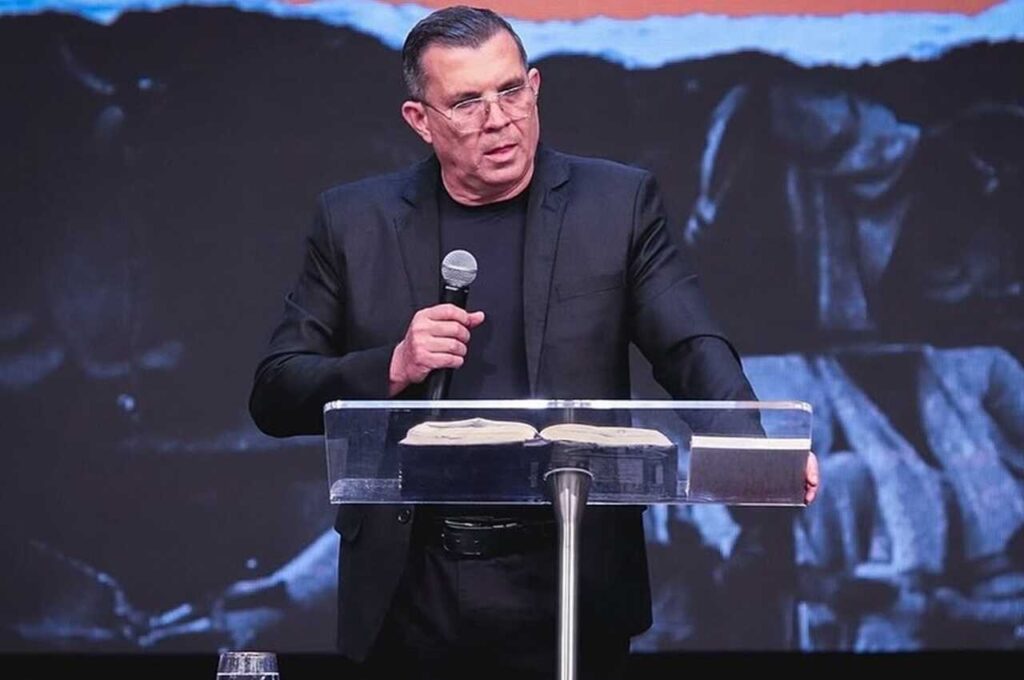 Pastor Cláudio Gama pregando ao microfone atrás do púlpito com a Bíblia aberta durante culto