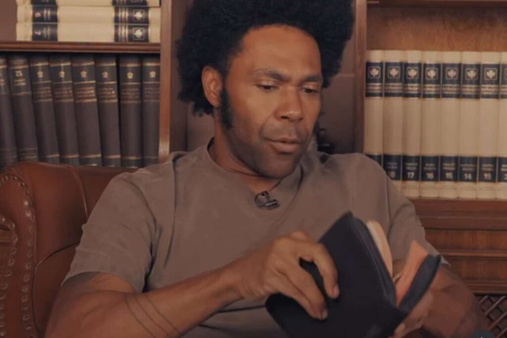 Cantor gospel Thalles Roberto manuseando uma Bíblia enquanto conversa durante gravação em estúdio com estante de livros ao fundo.