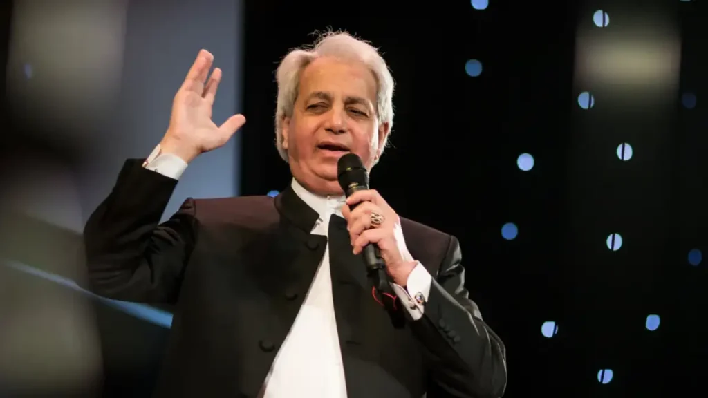 Televangelista Benny Hinn prega em palco segurando microfone e levantando a mão durante evento religioso