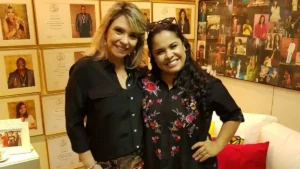 Cassiane e Marina de Oliveira posam juntas em estúdio com parede de quadros e fotos ao fundo