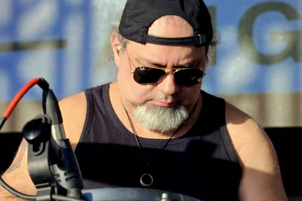 Baterista Geraldo Abdo tocando bateria durante apresentação, usando boné e óculos escuros.