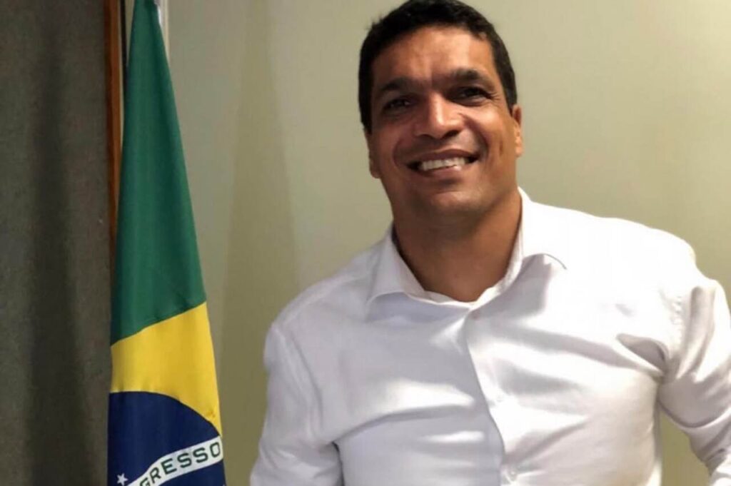 Cabo Daciolo sorrindo, vestindo camisa branca, com bandeira do Brasil ao fundo.
