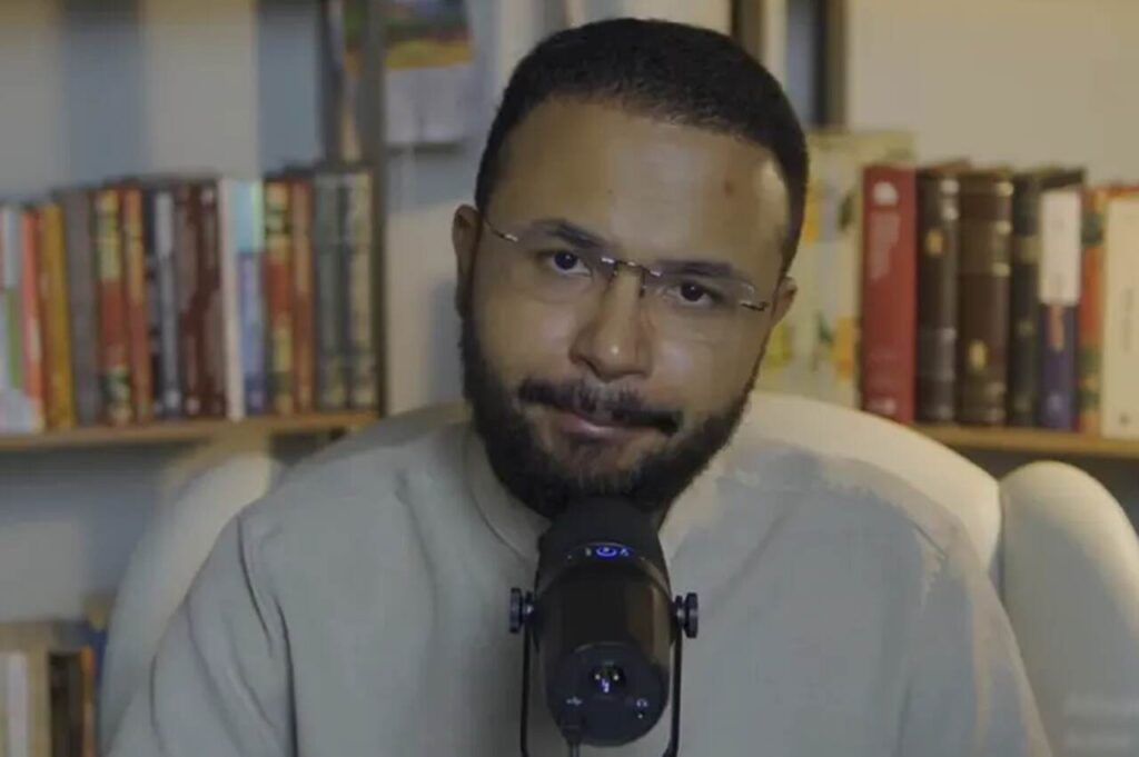 Caio Modesto fala ao microfone em estúdio, com estante de livros ao fundo.