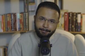 Caio Modesto fala ao microfone em estúdio, com estante de livros ao fundo.