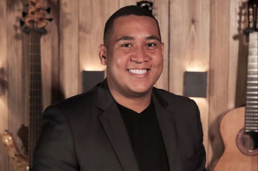 Cantor gospel Alisson Santos sorrindo em estúdio com instrumentos musicais ao fundo.