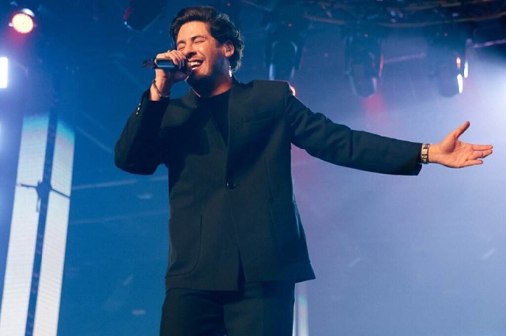 Cantor Marcos Freire se apresentando no palco, cantando com microfone e braços abertos.