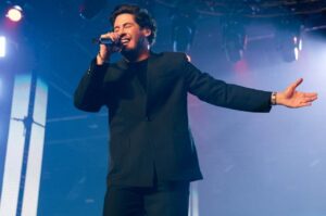 Cantor Marcos Freire se apresentando no palco, cantando com microfone e braços abertos.