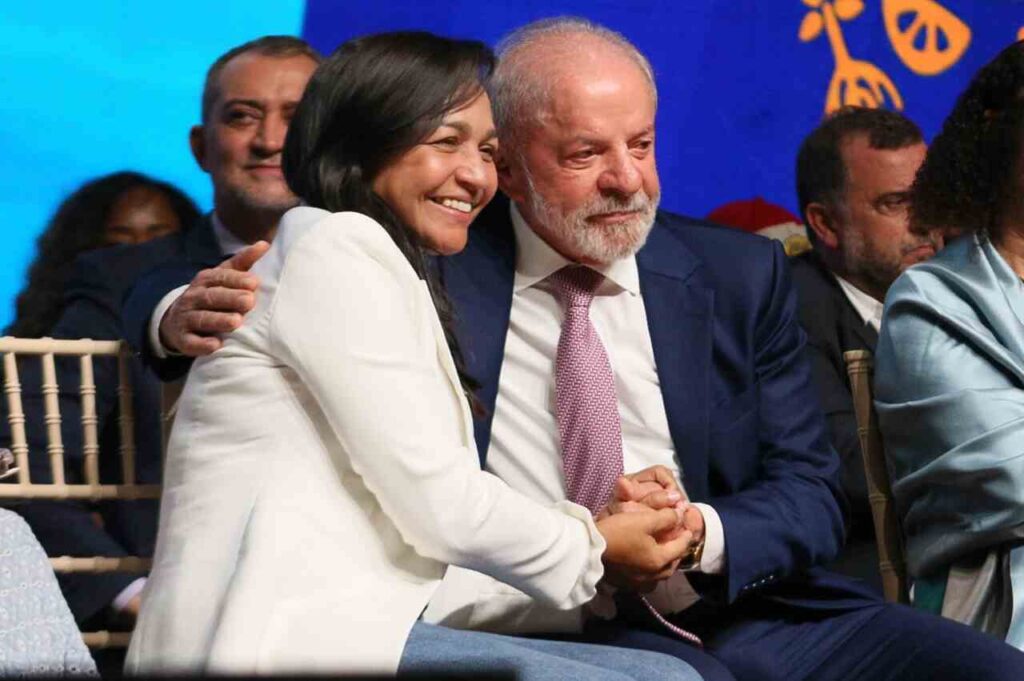 Eliziane Gama e Lula sentados lado a lado, de mãos dadas, durante evento público.