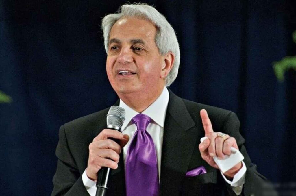 Pastor Benny Hinn pregando com microfone na mão, vestindo terno escuro e gravata roxa.