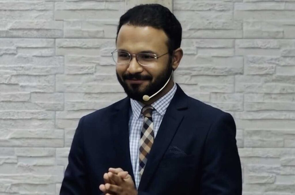 Pastor Caio Modesto pregando com microfone headset, vestindo terno e gravata, em ambiente de igreja.