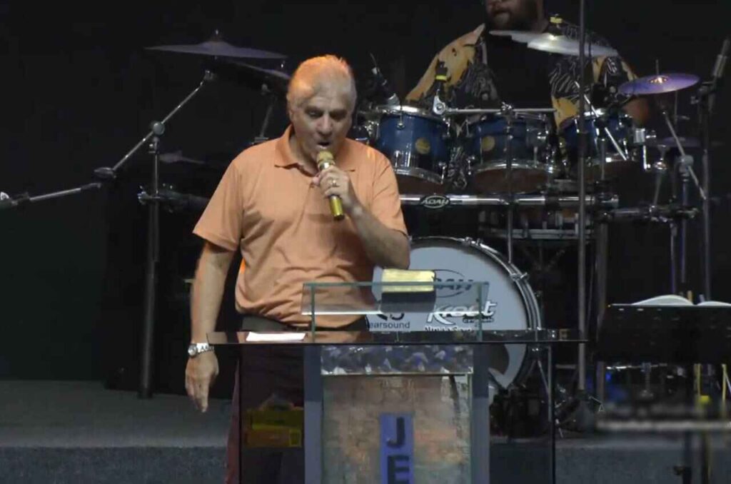 Pastor Carlos Moyses prega em púlpito durante culto, segurando microfone, com bateria ao fundo.