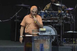Pastor Carlos Moyses prega em púlpito durante culto, segurando microfone, com bateria ao fundo.