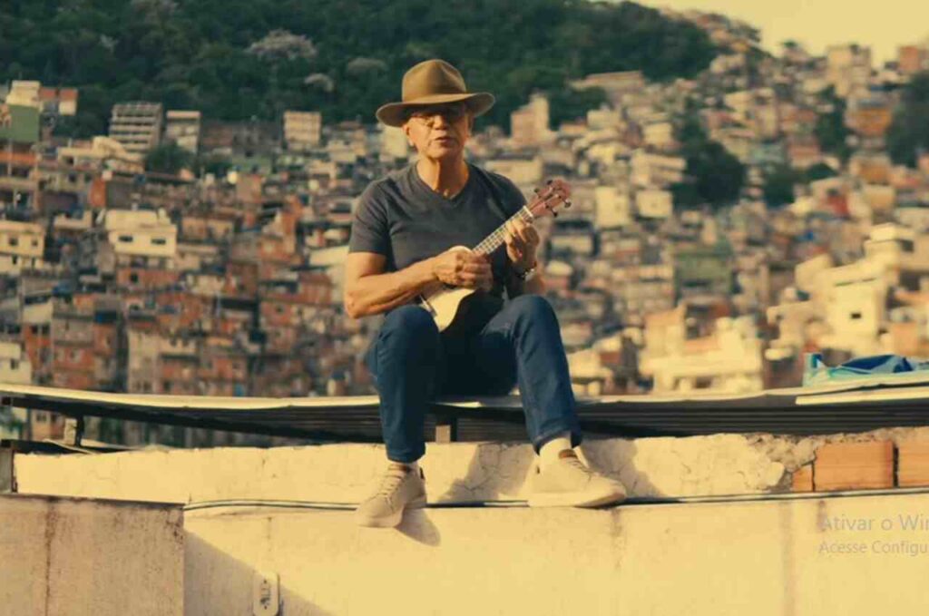 Sérgio Lopes toca instrumento de cordas sentado em laje, com comunidade ao fundo.
