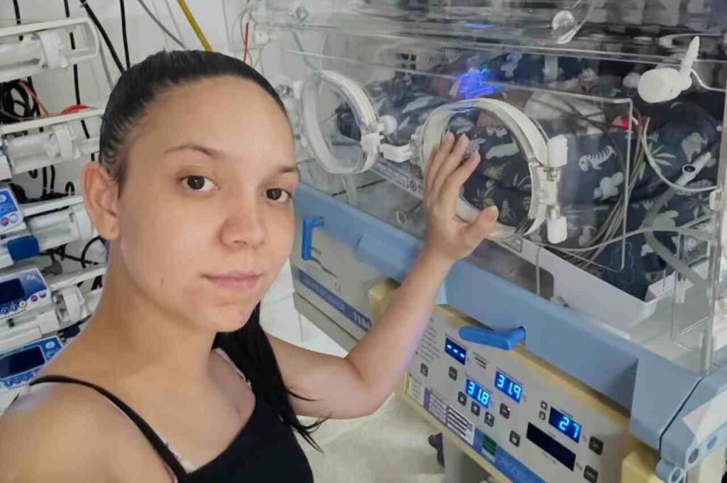 Mulher ao lado de incubadora neonatal em UTI, com bebê internado recebendo cuidados médicos.