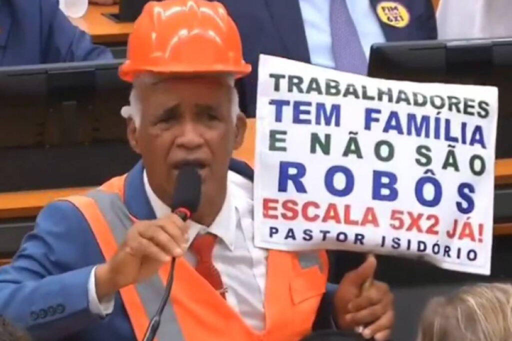 Deputado Pastor Isidório aparece usando capacete e colete refletivo enquanto segura cartaz sobre a escala 5x2 durante sessão legislativa.