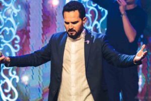Luciano Camargo aparece com os braços abertos durante momento de oração e adoração em evento cristão realizado em palco iluminado.